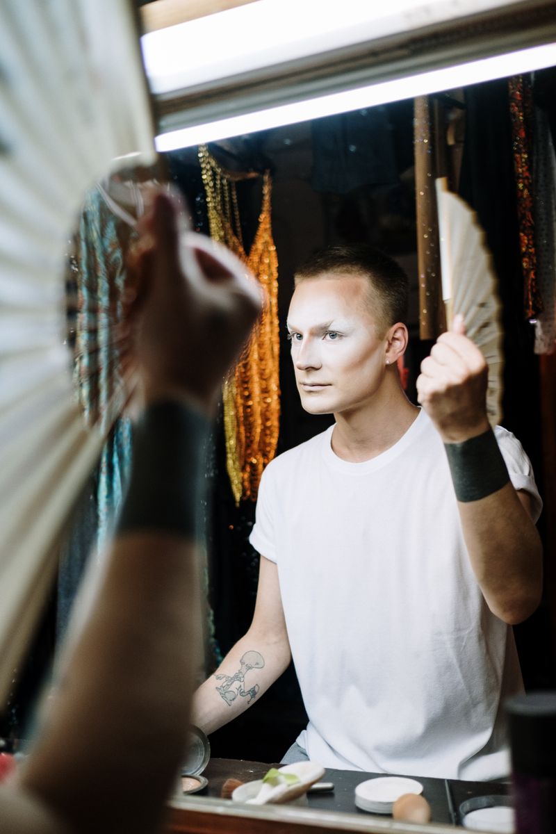 A drag performer applies makeup in a theatre dressing room, preparing for a performance.